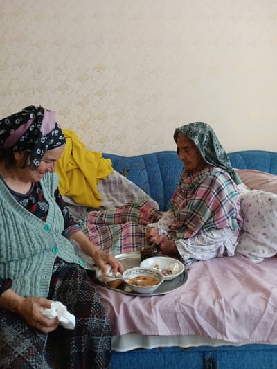 Two elderly women are seated on a couch in what appears to be a home. The woman on the left is wearing a light blue knitted cardigan over a patterned dress and headscarf. She is holding a white tissue in her left hand and reaching towards a food tray with her right. The woman on the right is wearing a patterned headscarf and a plaid shawl over a white floral dress. She is looking down at the food tray, which contains bowls of food.
The setting is a room with floral wallpaper. The couch is blue and is covered with various blankets and pillows. A yellow garment is draped over the couch. The lighting suggests it is daytime. The overall impression is one of a quiet, domestic scene, possibly a mealtime. The women's postures and expressions suggest a gentle, perhaps intimate moment. There is no visible text in the image.