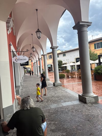 The image captures a scene at an outdoor shopping arcade in Barberino di Mugello, Italy, likely an outlet mall. The architecture features a series of white arches supported by substantial grey pillars, creating a covered walkway. The ground is paved with cobblestones and has a wet appearance, suggesting recent rain.

In the foreground, a man with long, gray, curly hair is seated, facing away from the camera, wearing a dark green t-shirt and denim shorts. To his right, a young boy in a yellow t-shirt and grey shorts walks towards the viewer, his gaze directed forward. Behind the boy, a woman in a black dress walks away from the camera, carrying a white bag. Further down the walkway, two figures are visible in silhouette, walking away.

Various signs are displayed on the buildings. One prominent oval sign reads "caffè MEDÍCEO" with small grey dots. Below it is a rectangular sign for "BREIL". In the background, more shops are visible, including one with a sign that appears to say "WOOLRICH". Umbrellas are open outside some establishments, and wooden benches are placed along the arcade. The sky is overcast with clouds.