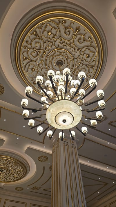 The image shows a view from below of a grand, ornate interior ceiling with a large chandelier. The ceiling features intricate gold-colored decorative moldings in a floral and swirling pattern. The chandelier itself is multi-tiered, with numerous lights pointing upwards and outwards, each encased in a white, cylindrical glass shade. The central part of the chandelier is a large, circular diffuser, also lit from within. To the right of the chandelier, a tall, fluted column with gold trim rises towards the ceiling, suggesting an elaborate architectural setting. The overall impression is one of opulence and grandeur, likely a ballroom, reception hall, or similar formal space. There are no people visible in the image. The location is stated as Agios Epiktitos, Northern Cyprus / Cyprus.