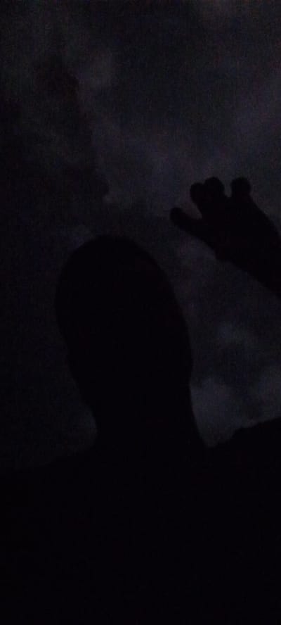 The image depicts a dimly lit scene, seemingly taken in Ilogbo Eremi, Nigeria. A dark silhouette of a person's head and upper body is visible against a background that appears to be a night sky or dark clouds. A hand, also silhouetted, is raised in the upper right portion of the image. The overall darkness obscures details, but the shapes suggest a possible self-portrait or candid shot captured in low-light conditions.