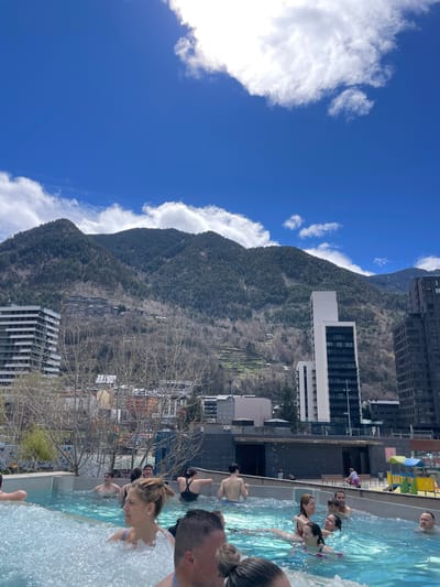The image captures a sunny scene in Les Escaldes, Andorra, likely on a rooftop pool. A group of people are in the pool, seemingly enjoying the water. Behind the pool, the mountainous landscape characteristic of Andorra dominates the view. There are some buildings, including tall residential ones, interspersed between the pools and the mountains. The sky is blue with scattered white clouds.