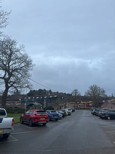 The image depicts an outdoor car park and commercial complex located at Durleighmarsh Farm, near Petersfield, East Hampshire, United Kingdom.
The sky is uniformly overcast and light grey, indicating an absence of direct sunlight. The ground surface of the car park is asphalt, appearing wet and reflective, suggesting recent precipitation.
In the foreground on the left, the rear of a silver pickup truck is partially visible, followed by a red Honda CR-V SUV (license plate LC22 EVR). Adjacent to it is a blue SUV (license plate YI70 RPF), and further vehicles, including dark blue, silver, black, and yellow cars, are parked in a row receding into the mid-ground. A tall, black lamppost with two illuminated white lights stands to the left of the red Honda CR-V. On the right side of the car park, a parallel row of parked vehicles is visible, including a light blue compact car in the distance.
To the left, a large, bare deciduous tree occupies the foreground, its branches extending into the upper left quadrant. In the mid-ground and background, additional bare deciduous trees are present, alongside dense evergreen foliage forming a treeline behind the buildings.
The background features a multi-story commercial building with a dark facade and numerous horizontal windows across its upper levels. Scaffolding is visible on the upper left portion of this building. A small, dark green canopy extends from a lower section of the building. To the right of this structure, a complex of traditional-style barn buildings with dark roofs and lighter-colored walls is visible, displaying internal and external illumination. A traditional red British telephone box stands in front of these barn buildings, positioned to the right of the central car park aisle. Small decorative lights are strung around the barn structures, and a line of small, wheeled objects is arranged nearby.
No individuals are visible, and no actions or interactions are depicted within the scene. The environment appears still and devoid of human activity.