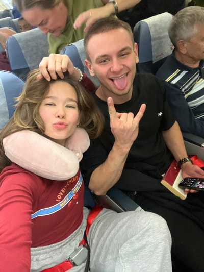 The scene is set inside an aircraft cabin, associated with Емельяново Airport, Russia. Two young adults are prominently featured in the foreground. A female with long, light brown hair, wearing a maroon long-sleeved shirt with "LOVELY CHERRY HAVE A GOOD DAY" text, light grey sweatpants, and a pink U-shaped neck pillow, is positioned on the left. She has her left hand placed atop her head, her lips pursed, and is secured by a visible red seatbelt. To her right, a male with short brown hair, wearing a black t-shirt with a white logo, black trousers, and a dark wristwatch on his left wrist, is seated. He extends his tongue and forms a "rock on" gesture with his right hand. His left hand holds a red-covered book with gold-edged pages and a black smartphone displaying app icons. In the row immediately behind them, a female with light brown hair and an olive-green top has her left hand reaching over the foreground female's head. Further back and to the right, an older male with grey hair and a striped shirt is visible, looking towards the right. Partial views of additional passengers are present in the background. The aircraft interior features blue and grey upholstered seats, each equipped with a white headrest cover, one bearing the text "SPACE".