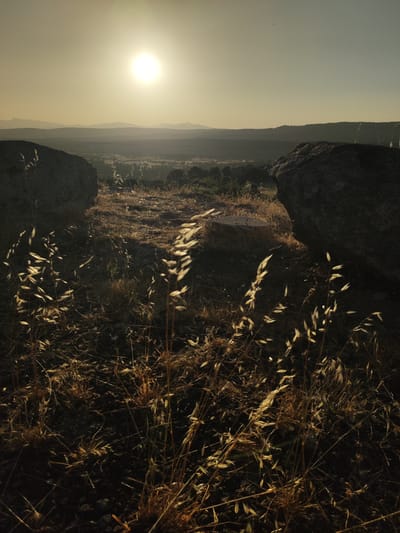 The image captures a serene landscape at sunset in Ledrada, Spain. In the foreground, dry grass and a few small stones are visible, bathed in the warm, golden light of the setting sun. Two large, dark rocks frame the scene, one on the left and another on the right, appearing to be part of the natural terrain. In the middle ground, the land slopes down to reveal a valley with scattered trees and agricultural fields. In the distance, a series of rolling hills and mountains recede into the horizon, softened by the atmospheric haze. The sun, a bright orb, hangs high in the pale sky, casting a gentle glow over the entire landscape. There are no people or animals present in the image, and the scene appears peaceful and undisturbed.