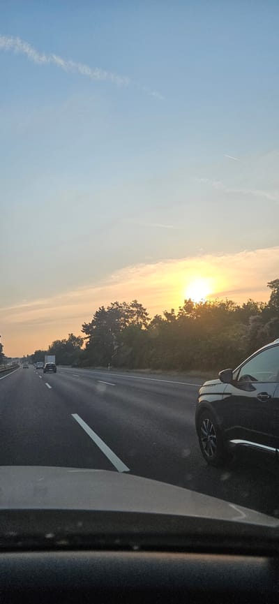 The image depicts a highway scene at sunset, likely on the M7 motorway approaching or leaving Székesfehérvár, Hungary, given the context of typical travel routes in that area. The sky is a gradient of pale blue to warm orange and yellow near the horizon, illuminated by the setting sun which is partially obscured by a line of trees. There is a contrail visible in the upper right portion of the sky. The highway itself is asphalt with white lane markings. In the foreground, the hood of a dark-colored vehicle is visible, as is the windshield. On the highway, several vehicles are in motion: a black SUV is in the right lane, and further ahead, a white truck and other cars are visible in the left and middle lanes. The vegetation along the side of the highway consists of dense trees and bushes. The overall impression is of a journey during dusk.