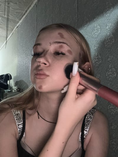 The image shows a young woman with blonde hair, applying makeup. She has several brown spots on her face, which may be part of the makeup application or a skin condition. She is wearing a black top with sparkly straps. A makeup brush is being used on her cheek. The background appears to be a textured wallpaper. There is no information in the image that indicates the location is in Velykyi Sukhodil, Ukraine.