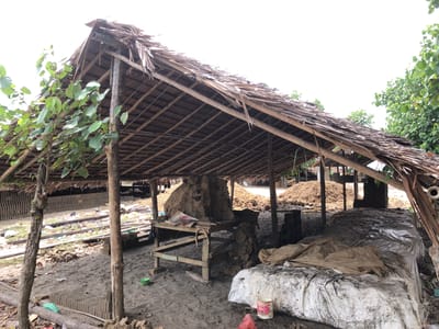 The image depicts an open-air structure with a thatched roof, likely located in a rural or industrial area, possibly a brick-making facility, in Bangkajaya, Indonesia. The structure is supported by wooden poles and beams, with the roof made of dried palm leaves or similar vegetation. The ground is sandy and uneven, with piles of soil or clay visible in the background, suggesting raw materials for brick production. There are stacks of what appear to be unfinished bricks in the midground and background, some covered with plastic sheeting. A rustic wooden table or workbench is present under the shelter, with some tools or materials on it. The overall scene is one of manual labor and production in a natural environment.