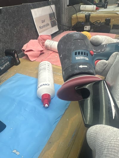 In an indoor workspace located in Fulda, Germany, a gloved individual is actively polishing a black, reflective, curved object. The operator's right hand, clad in a grey knit-textured glove, holds a blue and black Bosch cordless power tool, identified by "BRUSHLESS MOTOR" text and a visible 12V battery pack. A reddish-brown polishing pad is attached to the tool, which is in motion.
To the left of the polishing activity, a white plastic bottle with a red flip-top cap, labeled "ONSYST" and indicating "CUT 6/10" and "GLOSS 8/10," stands upright on a blue protective mat. White residue is present on the bottle's nozzle. The blue mat, which is slightly crumpled, rests on a yellowish-brown workbench surface and exhibits scattered white specks.
In the background, a white document within a clear plastic sleeve displays the German text "zur Kontrolle" (for inspection) and is mounted on a dark, granular-textured wall. A crumpled pink microfiber cloth lies nearby. Further back, two black electronic devices, likely battery chargers, are visible with connecting cables on an elevated surface; one displays a blue indicator light. A separate 12V cordless drill battery is positioned on the left side of the frame.