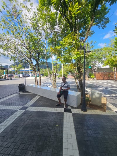 The image depicts a man sitting on a modern, angular bench outdoors in a paved area. He is wearing a grey and white striped t-shirt and dark shorts, with sandals on his feet. The bench is made of white marble with grey veins and has a sharp, geometric design. The man appears relaxed, looking to his left.

The setting is a pedestrian walkway or plaza with a distinctive patterned pavement. The pavement consists of dark grey tiles arranged in a grid, with alternating white tiles forming diagonal stripes and crossing patterns. The bench is positioned alongside this tiled area.

Behind the bench, trees with lush green foliage and some small, spherical lights are visible, suggesting a park-like or well-landscaped urban environment. In the background, more trees line a road, with a building that has wooden fencing and white walls. Further in the distance, mountains are visible under a bright blue sky with scattered white clouds, indicating clear weather. The presence of a gas station with cars and signage in the background provides a further cue to the location. The lighting suggests it is daytime, likely late morning or early afternoon, given the strong shadows. The overall atmosphere is peaceful and casual.