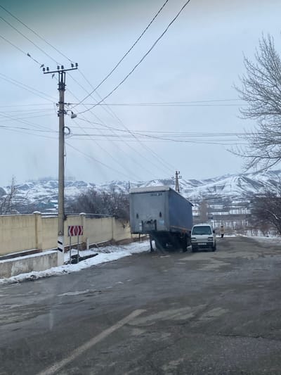 The image depicts a winter street scene in Norayburi, Uzbekistan, under an overcast grey sky. Patches of snow and slush are present on the ground alongside a worn asphalt road. A light-colored concrete wall runs along the left side of the road, with a white and black striped road marker displaying a double red chevron (>>) attached to a smaller post. A tall concrete utility pole stands in the mid-ground left, supporting numerous overhead power lines and a street lamp; several small birds are perched on its upper wires. To the right, a large grey and blue semi-trailer, bearing the labels "AKHTAN" and "TUR", is parked on the roadside, detached from its tractor and resting on its landing gear. A white small van, with the license plate H 801 GB, is parked directly in front of the semi-trailer. A single individual, dressed in a light-colored coat, is visible further down the road on the right, standing behind the white van and facing away from the camera. The background consists of snow-covered hills and mountains, sparsely populated with bare winter trees.