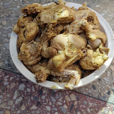 A white, circular bowl is filled with numerous pieces of cooked, seasoned chicken. The chicken, primarily comprising legs and various body cuts, exhibits a yellowish-brown hue with visible seasoning residues and some skin. The bowl is positioned on a multi-colored terrazzo floor, characterized by irregular fragments of reddish-brown, grey, and off-white aggregate set within a lighter cementitious matrix. The scene is brightly illuminated by ambient light, suggesting an outdoor or well-lit indoor environment in Dan Maliki, Nigeria. No individuals or additional objects are visible within the frame.