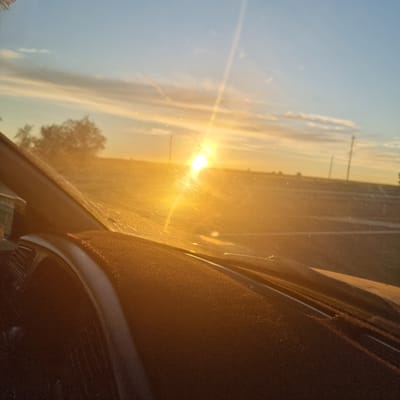 The image shows a sunset viewed through the windshield of a vehicle. The sun is a bright, glowing orb on the left side of the frame, casting a warm, golden light across the scene. Streaks of clouds are visible in the sky, tinged with orange and yellow hues. The landscape outside the car appears flat and open, with a faint suggestion of distant trees on the right and power lines in the distance. A portion of the car's interior, including the dashboard, is visible in the lower right corner, in shadow. The overall impression is one of a serene and peaceful moment during a drive at dusk. The time of day is clearly sunset due to the low angle of the sun and the warm, diffused light. The weather appears clear with some scattered clouds. The location context suggests the scene is in Alğa, Kazakhstan. There is no visible text in the image.