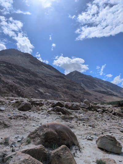This is a vertical, eye-level, medium shot of a rocky, mountainous landscape under a bright, cloudy sky. The foreground is dominated by a field of various-sized rocks, from pebbles to large boulders, scattered across a sandy and dusty ground. In the mid-ground, a steep, rocky mountainside rises sharply, its textured surface showing layers of sediment and loose stones. Further in the background, another mountain peak is visible, appearing further away and less detailed. The sky is a vibrant blue, dotted with fluffy white clouds, with the sun's glare visible in the upper left quadrant. A hint of green vegetation can be seen on the far right, at the base of the mountains. The scene appears to be an arid, natural environment, likely a desert or mountainous region. The location is identified as Sasoma, India. There are no people or animals present in the image.