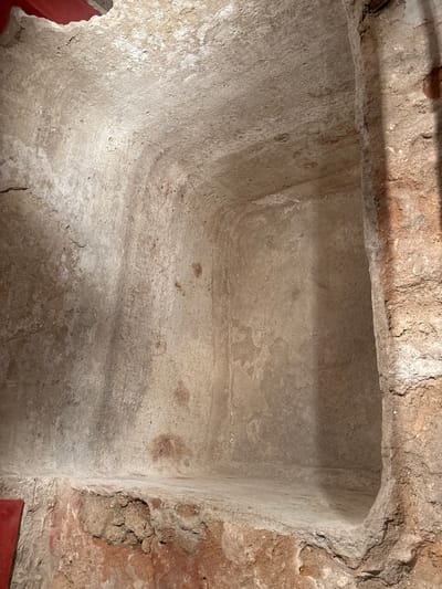An empty, ancient, rectangular burial niche is depicted within the Phoenician Necropolis in Cádiz, Spain. The niche, roughly carved into rock or earth, presents a flat base, vertical side walls, and a flat ceiling. All interior surfaces are coated in a light-toned, eroded plaster or stucco, exhibiting extensive calcification, textural variations, and multiple irregular brown and reddish-brown discolorations consistent with mineral staining and age. The adjacent wall to the right shows a rougher, unrefined excavation surface. A small, dark red, rectangular object, possibly modern interpretive signage, is partially visible in the lower left foreground. Artificial illumination highlights the textures and depth of the recession. No human figures or specific artifacts are present within the niche.