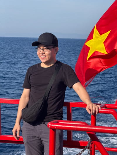 A person wearing a black t-shirt, grey pants, and a black baseball cap stands on a red railing by the sea. They are smiling and looking to the left. A Vietnamese flag is visible to the right of the person. The sea is blue and calm with a clear sky. The railing appears to be part of a boat or ferry. The image does not provide any information to suggest a location in Dong Son, Vietnam.