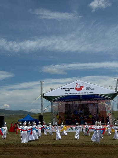 This photograph captures an outdoor cultural performance in Orongoi, Russia. A large, white tent-like structure with signage in Cyrillic script covers a stage. The stage features a screen displaying a colorful, animated scene with traditional figures and banners. On the stage itself, several individuals in traditional dark attire appear to be performing.
In front of the stage, a group of approximately ten people, primarily women, are dressed in white robes and conical hats, holding and maneuvering long, brightly colored sashes of red, blue, green, and yellow. Their movements suggest a choreographed dance. To the left of the main performance area, a smaller group of people in red and white attire are visible, and further left, a blue tent is set up.
The ground is covered in dry grass, with a patch of dark, tilled earth in the foreground. In the background, rolling hills with sparse vegetation and scattered trees extend towards a hazy horizon. The sky is a vibrant blue with scattered white clouds, some wispy and others more substantial. The overall scene is one of a festive outdoor gathering celebrating cultural traditions under a bright, partly cloudy sky.