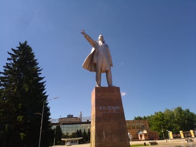 In the city of Otradny, Russia, a full-body bronze statue of Vladimir Lenin stands prominently on a tall, square, copper-colored pedestal. Lenin is depicted in a long coat, with his left arm outstretched and his index finger pointing upwards, as if addressing a crowd or making a proclamation. The pedestal is inscribed with his name, "В.И. ЛЕНИН", and his birth and death dates: "22.04.1870 21.01.1924".
Behind the statue, a large, modern building with a glass facade and the word "РОССИЯ" (Russia) in large letters is visible. To the left of the statue, a tall, dark green evergreen tree stands, partially obscuring the view of the sky. Streetlights and some smaller buildings are also present in the background. The sky is a clear, bright blue with a few wisps of clouds. The overall scene suggests a public square or plaza in the city.