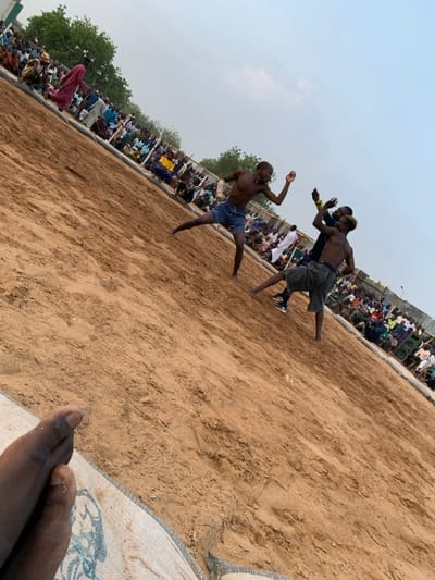 The image shows three shirtless men wrestling in a sandy area. Two are grappling, while the third is kicking one of the grapplers in the air. The men are surrounded by a large crowd of spectators, who are seated on the ground. The area is dry and dusty, with little vegetation visible. The coordinates provided place this scene in the city of Sokoto, Nigeria. The image is shot from a low angle, with the subject in the foreground being a person's hand. The sand is a light brown color and appears to be a natural sand pit in a public or semi-public area. There are many spectators in the background wearing various styles of clothing, including some white robes. This is likely a traditional wrestling match taking place in Sokoto, Nigeria.