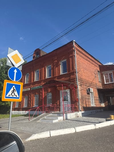 In the city of Vereya, Russia, a brick building with a red facade stands under a bright blue sky. The building, which appears to be a commercial establishment with the text "АПТЕКА" (Pharmacy) visible above the entrance, features multiple windows with metal grates and a red awning over the doorway. A ramp and stairs lead up to the entrance, bordered by a red metal railing.
In the foreground, a traffic signpost displays three signs: a diamond-shaped sign with a yellow center and white border, a circular blue sign with a white upward-pointing arrow, and a square blue and orange sign with a white pedestrian crossing symbol. The ground is paved with gray interlocking bricks, and a portion of a car's side mirror is visible in the bottom left corner. Overhead, black electrical wires are strung across the sky. The scene is bathed in sunlight, casting shadows on the pavement.