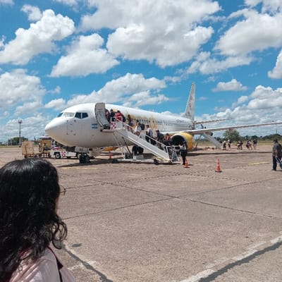 An airplane, predominantly white with yellow accents, is parked on the tarmac with passengers descending the stairs. A woman with dark, curly hair is visible in the foreground. The sky above is a vibrant blue with scattered, fluffy white clouds. Other individuals are seen further down the tarmac. The ground is concrete.