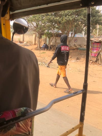 An image captured from within a vehicle, likely a Keke Napep (tricycle), shows a street scene in Goron Dutse, Nigeria, under bright daylight. In the foreground, the back of a person wearing a dark brown garment is visible on the left, with a circular side-view mirror reflecting a blurred exterior above their left shoulder. A shiny metal handrail curves horizontally across the lower central portion of the frame.
In the midground, a young male walks from left to right on a reddish-brown dirt path. He wears a black hooded sweatshirt with red and white text on the back, including large red characters (appearing as "后庄") and English text that reads "IT IS MY PRIDE." He is also wearing orange shorts and light-colored flip-flops. To the left of the walking individual, a group of three men are seated on a low structure or ground under trees, conversing or resting, facing away from the camera. Behind them, a light-colored concrete wall is visible.
The background features multiple trees with green foliage. To the far right, a rudimentary structure with a corrugated metal roof and a wall partially covered by colorful, checkered fabric (red, pink, white) is present. The ground throughout the visible area is dry, reddish-brown earth.