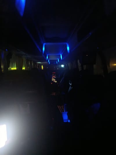 The image shows the interior of a vehicle, possibly a bus or train, illuminated by blue LED lights. The lighting is dim, creating a dark and atmospheric setting. In the foreground, on the left, a bright white light source is visible, possibly a camera flash or a phone screen. The interior appears to be empty of people in the immediate foreground, but silhouettes of passengers can be seen further back in the vehicle. The overhead compartments and lighting fixtures are visible. The image lacks any specific indicators of location within the city of Maiaky, Ukraine, or any identifiable landmarks. The scene is characterized by its low-light conditions and the presence of blue accent lighting.