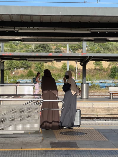 The image depicts a scene at a train station, likely in Granollers, Spain, on a sunny day. Two women in traditional Islamic attire are standing on a platform, facing away from the camera. The woman on the left wears a dark brown abaya, while the woman on the right is dressed in a light beige hijab and a long grey dress, holding a small silver suitcase. A third person, a young woman with a blue backpack and pink pants, is visible in the background walking away from the two women. The platform is made of concrete and textured tiles, with yellow tactile paving along the edge. A metal railing and a perforated metal screen protect a set of stairs to the left. In the background, there is a railway track, a bench, a trash can, and a hillside covered with trees and vegetation. The station appears to be covered by a metal canopy with fluorescent lighting.