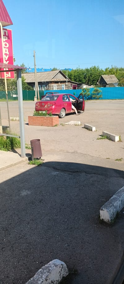 A red sedan is parked on a dusty asphalt lot. The driver's side door is open and a person's legs, wearing white shorts and black sandals, are visible exiting the vehicle. A planter box filled with greenery sits in front of the car. To the left of the planter, a rusty metal trash can is positioned near a bus stop shelter. The background shows a wooden house with a blue metal roof, a blue fence, and green trees under a clear blue sky. The scene appears to be in the village of Kiyasovo, Russia, as indicated by the Cyrillic text on a red sign visible on the left side of the image.