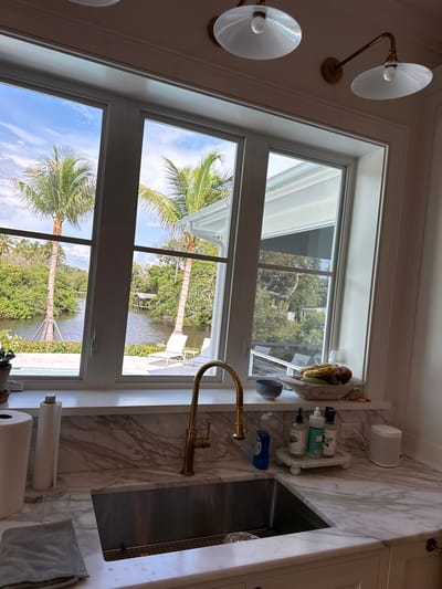 The image depicts an indoor view of a kitchen counter and sink area, looking out through a large window. The window offers a scenic view of a tropical waterway with palm trees and lush vegetation. In the foreground, the kitchen features a marble countertop with a stainless steel sink, a gold faucet, a roll of paper towels, a gray cloth, and several bottles of soap. A fruit bowl containing bananas, mangoes, and apples sits on a shelf above the sink. In the background, visible through the window, are two white lounge chairs and a small covered dock on the water. The ceiling above the window has two light fixtures. The scene appears to be in East Naples, Florida, United States of America, given the tropical vegetation.
