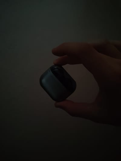 A person's hand is holding a small, dark gray, rectangular object with rounded edges, which appears to be a charging case for wireless earbuds. The background is a plain, dimly lit wall. The image was taken in Sosnowiec, Poland.