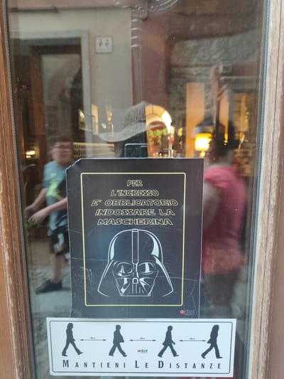 The image shows a glass-paneled door with a sign featuring Darth Vader and social distancing icons. The sign, in Italian, reads "PER L'INGRESSO E' OBBLIGATORIO INDOSSARE LA MASCHERINA" which translates to "For entry, it is mandatory to wear a mask." Below this text is an illustration of Darth Vader's helmet. Further down, a rectangular sticker displays silhouettes of people walking, with arrows indicating a distance of "1 Metro" (1 meter) between them. The text below this sticker says "MANTIENI LE DISTANZE" which means "Keep your distance."

The reflections in the glass show a blurred interior of a shop or establishment. A person wearing glasses and a t-shirt is visible on the left, appearing to be inside the building. On the right, the blurred reflection of a person in a red top is also visible. A straw hat is reflected on the top right of the glass. The door frame is made of dark wood.

The location is identified as Orta San Giulio, Italy. The overall scene depicts a COVID-19 safety measure implemented at an entrance.
