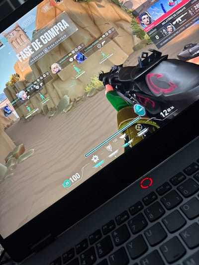 The image shows a laptop screen displaying the game Valorant. The in-game phase is the "Buy Phase" where players can purchase weapons and abilities. Agent information and combat reports are visible. A masked character is present in the game. The laptop's keyboard is partially visible, including the power button with a red ring. The game appears to be played on a desert-like map with a sandy texture.
