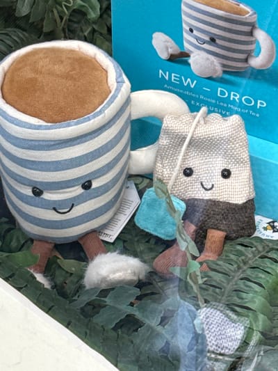 The image displays a retail window exhibit in Ambleside, United Kingdom, featuring two plush toys from the "Amuseables Rosie Lea Mug of Tea" collection by Jellycat. The primary subject on the left is a stuffed tea mug, characterized by white and blue horizontal stripes, a light brown interior representing tea, a white handle, and a smiling anthropomorphic face with black eyes and a black stitched mouth. It has four small brown legs ending in white, fluffy feet, and a white product tag is visible on its right side. To the right of the mug, a second plush toy depicts a tea bag, segmented into a light beige, textured upper portion and a dark brown lower portion. This tea bag also features a smiling anthropomorphic face with black eyes and a black stitched mouth, along with four small brown legs. A white string extends from its top, adorned with a small teal fabric square and a green fabric leaf. Both plush toys are positioned on a bed of artificial green foliage, resembling ferns. The background consists of a bright blue surface displaying white text that reads "NEW - DROP" and "Amuseables Rosie Lea Mug of Tea EXCLUSIVE," with a graphic depiction of the tea mug toy. Reflections from the external environment are visible across the foreground, indicating a glass barrier. No people are present.