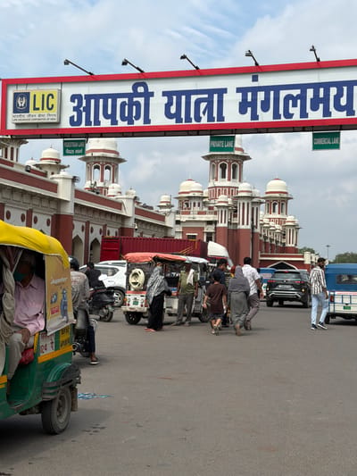The image shows a street scene in front of a red and white building with multiple domes and arches, characteristic of Indian architecture. A large banner with the text "LIC आपकी यात्रा मंगलमय" (LIC Your journey is auspicious) is prominently displayed overhead, indicating the presence of the Life Insurance Corporation of India. Several vehicles, including auto-rickshaws, a car, and a van, are on the road. Pedestrians are walking in various directions. Signs for "PEDESTRIAN LANE," "PRIVATE LANE," and "COMMERCIAL LANE" are visible, suggesting traffic management within the area. The overall environment appears to be a busy public space in the city of Lucknow, India.