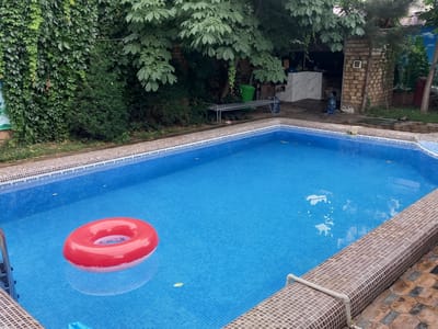 This is an outdoor shot of a swimming pool in Charvak, Uzbekistan. The pool is filled with bright blue water, and a red inflatable ring floats on the surface. The pool is surrounded by a tiled border, and beyond that, the ground is paved with stone tiles. Lush green foliage and trees surround the pool area, and in the background, there is a brick structure with an outdoor kitchen or barbecue area. The overall impression is of a relaxed, inviting recreational space in a natural setting.