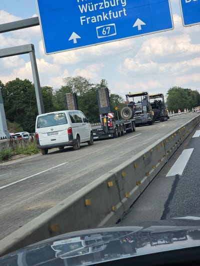 The image shows a section of a German autobahn with traffic cones and road construction equipment. In the foreground, the hood of a car is visible, reflecting the scene. To the left, a white Volkswagen Transporter van is parked on the side of the road. Further ahead, two large pieces of road construction machinery, likely asphalt pavers, are positioned on the roadway. A large trailer with a spare tire is attached to one of the pavers. A blue highway sign with white lettering indicates directions to Würzburg, Frankfurt, and the number 67. The environment is characterized by a cloudy sky and green trees lining the roadside. The scene suggests ongoing road maintenance or construction. The provided information about Rüsselsheim am Main, Germany, is not directly evident from the visual content of the image itself, but the presence of German highway signage and vehicle license plates with German formatting strongly suggests a German location.