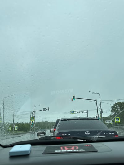 The image shows a view from inside a car looking out at a wet street in Пионерский, Russia. The windshield has water droplets on it, indicating rain. Traffic lights are visible, with one showing a green light. There are several cars on the road, including a dark-colored Lexus LS 430 in the foreground. Pedestrian crossing signs are also present. The sky is overcast and grey. The overall scene depicts a rainy day in an urban or suburban environment.