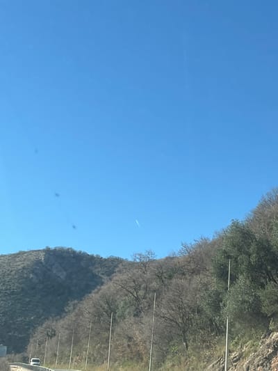 A panoramic landscape photograph, reportedly captured in Brežine, Montenegro, depicts a mountainous road scene under a clear sky. The upper two-thirds of the frame are dominated by a cloudless, light blue sky. Several dark, irregular smudges are visible in the upper left quadrant, consistent with dust or imperfections on a camera lens or viewing surface. A faint, white contrail, indicative of an aircraft's passage, is present in the upper right, arcing subtly downwards.
The lower third of the image features rugged hills and mountains, their slopes covered with sparse vegetation. Numerous trees appear deciduous and barren, while some sections exhibit green evergreen foliage, particularly on the right side. Along the base of the mountains, a paved road is visible in the lower left portion of the image. A white vehicle, possibly a van or car, is partially visible on this road, moving away from the viewer. Multiple slender, light-colored poles, consistent with utility or street light poles, are evenly spaced along the roadside, extending vertically into the midground from the lower center and right. No individuals or direct interactions are discernible within the scene.