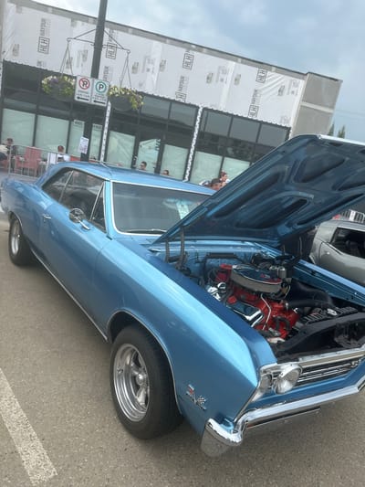 This media file depicts a light blue classic car with its hood open, showcasing its engine. The car appears to be a Chevrolet Chevelle SS, identifiable by the badges on the fender and the overall body style. The scene is set outdoors on what appears to be a street or parking lot, with a building under construction in the background. The building is covered in white sheathing with "HLH HAYDUK LUMBER AND HARDWARE" printed repeatedly. Visible signage on a pole indicates parking regulations, including "EVERYDAY 2 A.M.-7 A.M." and "EVERYDAY 9 A.M.-5 P.M. 2 HOUR PARKING". The sky is overcast, suggesting a cloudy day, and the time of day is indeterminate. Several people are visible in the background, some near the building entrance, others near the car, suggesting a car show or public gathering. The overall mood is casual, with an emphasis on the vintage automobile.
