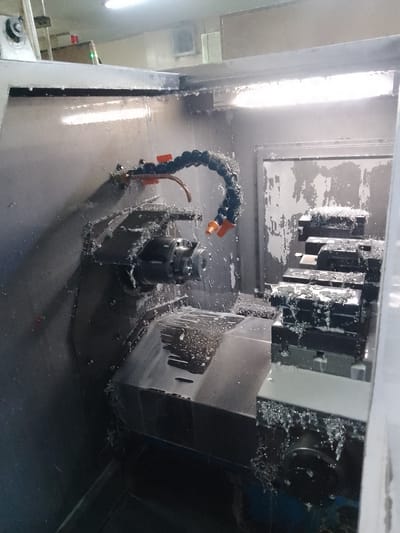 The image shows the interior of a CNC machine in a workshop. The machine is actively cutting metal, evidenced by the spray of coolant and metal shavings. A flexible hose with an orange nozzle directs coolant onto the workpiece. In the foreground, a chuck is visible on the left, and a stack of workpieces held in vises is on the right. Metal shavings are accumulated on various surfaces within the machine. The environment appears industrial, with fluorescent lighting overhead. The image does not provide any specific information about the city of Ciantra, Indonesia, or any people present.