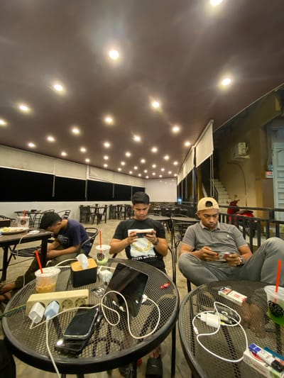 The horizontally oriented image depicts a nighttime scene at an outdoor cafe or terrace in Keudeublang, Indonesia, featuring three young men gathered around two metal tables. The area is illuminated by numerous bright, warm-toned string lights or spotlights on the overhead awning or ceiling.
In the foreground, a man wearing a gray polo shirt and a light-colored baseball cap looks towards the camera, holding a smartphone. On the table next to him are a pack of "CLASSIC" cigarettes, a "BLANC" lighter, a drink in a clear plastic cup with an "88" logo and a red straw, along with charging cables and a white power adapter.
At the adjacent table, a man in a black t-shirt, featuring a partially visible graphic that reads "COEN", holds a phone, looking down. Another man, wearing a blue t-shirt, is seated beside him, leaning forward with his head down. This table is laden with multiple smartphones, a black laptop or tablet, white earbuds, and a power strip connected to various chargers. Another drink with an "88" logo is visible, alongside a small item labeled "MEJA".
The background reveals more tables and chairs, a staircase, and a red scooter or motorcycle parked near a building. The atmosphere suggests a casual social gathering where the individuals are engaged with their electronic devices, utilizing the accessible charging facilities.