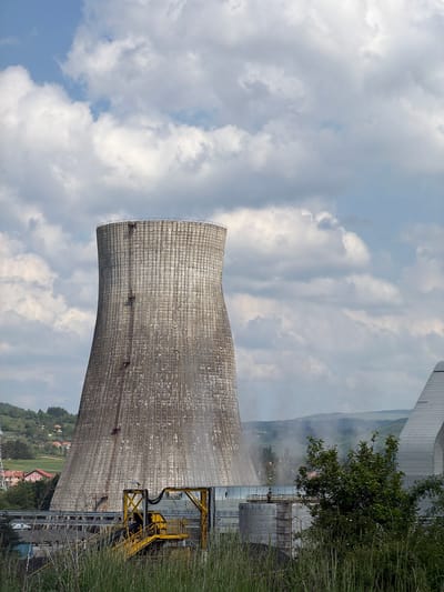 The image shows a cooling tower, likely part of a power plant or industrial facility. The tower is cylindrical with a flared top, constructed from concrete or a similar material. It is located in the city of Zabrđe, Montenegro.
The sky above the tower is filled with clouds, suggesting a partly cloudy day. To the side of the tower, there is some vegetation, including grass and trees. A structure with yellow supports can be seen adjacent to the tower. In the background, there are hills or mountains with some settlements visible. There is no apparent sign of any people or actions.