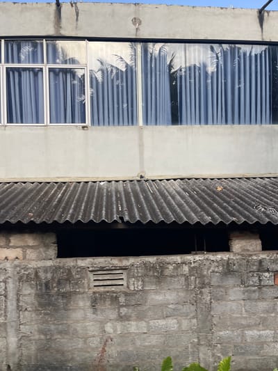 The image shows a close-up view of a two-story building, likely in Maggona, Sri Lanka, focusing on the facade. The upper story features a large window with blue curtains and a reflection of palm trees. The lower story has a corrugated metal roof supported by brickwork, and a rough concrete wall with a ventilation grille. The scene appears to be an exterior view, possibly a residential or commercial structure. There are no people or discernible actions visible in the image. The environment is a building exterior, with a clear sky visible in the top portion of the frame. The overall impression is one of a simple, functional building.