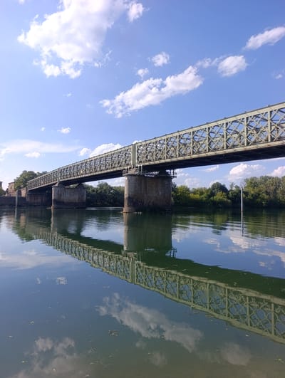 The image depicts a steel truss bridge spanning a body of water, likely a river, under a bright blue sky with scattered white clouds. The bridge is supported by stone pillars. The water reflects the bridge and the sky, with some debris floating on its surface. Lush green trees line the far bank of the river. The scene is viewed from a low angle, close to the water's surface. This bridge is located in Guéreins, France.