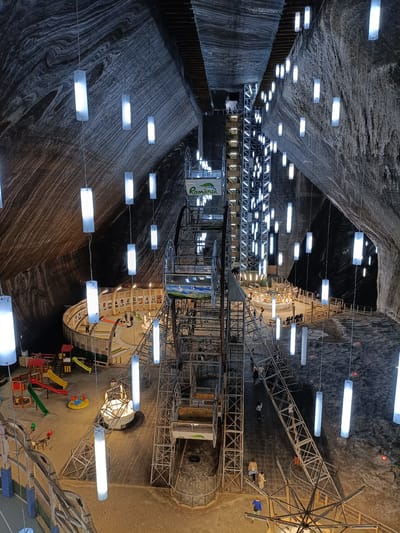 An eye-level, high-angle shot captures the interior of the Salina Turda salt mine in Turda, Romania, transformed into an amusement park. The cavernous space is characterized by rough, textured salt walls and a high ceiling, illuminated by numerous hanging, rectangular white lights and vertical strip lights on a central, multi-story metal structure. In the foreground, a section of a playground is visible, featuring a red and yellow climbing structure with slides and a yellow merry-go-round. Further into the mine, a circular wooden walkway encircles a central attraction that appears to be a small Ferris wheel or a similar ride. People are scattered throughout the scene, some walking on the walkways, others gathered near the attractions. A prominent sign with the word "România" and a green leaf logo is attached to the central structure. The overall impression is that of a surreal, subterranean entertainment venue carved out of a natural salt formation.