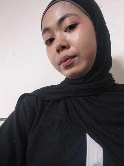A selfie, taken indoors against a plain white wall, shows a young woman wearing a black hijab and a black ribbed long-sleeved shirt. Her face is turned slightly towards the camera, and she is wearing makeup, including pink blush on her cheeks and eyelids, and a pinkish-brown lipstick. Her eyes are open and looking at the camera. The image is a close-up, focusing on her face and upper torso. There are no other individuals or objects clearly visible in the frame. The lighting is artificial and somewhat harsh, creating reflections on her skin. The background is a plain, undecorated wall. The location in Jeppee, Indonesia, is not discernible from the image itself.