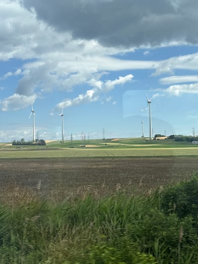The image shows a rural landscape with several wind turbines in the background. The foreground consists of fields of crops, with tall green grass and reeds in the immediate foreground. The sky is partly cloudy with a mix of blue sky and white and gray clouds. The wind turbines are white with red tips on their blades and are situated on rolling hills. Power lines and transmission towers are also visible across the landscape. The scene appears to be a sunny day, given the brightness of the sky and the illumination of the landscape.