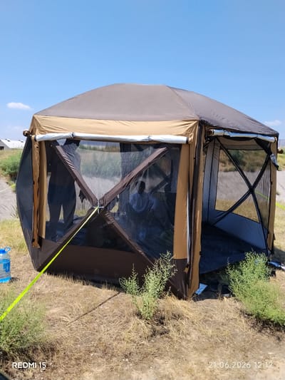 An outdoor scene captured on a sunny day in Yovon, Tajikistan, features a tan and brown pop-up screened canopy tent with a dark roof. The tent has mesh walls that allow visibility into the interior, where two figures, silhouetted and appearing to be adults, are seated. A blue plastic water bottle sits to the left of the tent. The surrounding environment consists of dry, yellowed grass and scattered green bushes. In the background, a dirt road is visible, leading towards distant buildings and a clear blue sky. The timestamp "21.06.2026 12:12" and "REDMI 15" are overlaid on the bottom of the image.