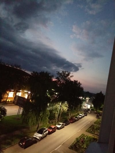 The image captures a dusk scene in a city, possibly Alatyr, Russia, with a dramatic cloudy sky transitioning from dark gray to lighter hues with hints of pink near the horizon. In the foreground, a street is lined with parked cars. A white SUV with black trim is prominent, followed by a red sedan and other vehicles. To the left of the street, a grassy area separates the road from buildings. Trees with dense foliage obscure some of the buildings, but illuminated windows suggest occupancy. A large billboard with the word "HOOKA" is visible, leaning on its support structure. The overall atmosphere is calm, with the natural light fading and artificial lights beginning to illuminate the area.