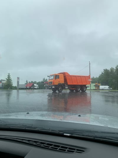 An orange dump truck is parked in an outdoor lot on a cloudy day. The ground is wet, suggesting it has rained recently. In the background, there are several buildings and trees. A white van is parked to the right of the dump truck. The image appears to be taken from inside a vehicle, as the dashboard and windshield are visible in the foreground. The scene is likely a truck stop or a service area. The image is taken in the vicinity of Yunkoŭski sel'ski Saveč, Belarus.